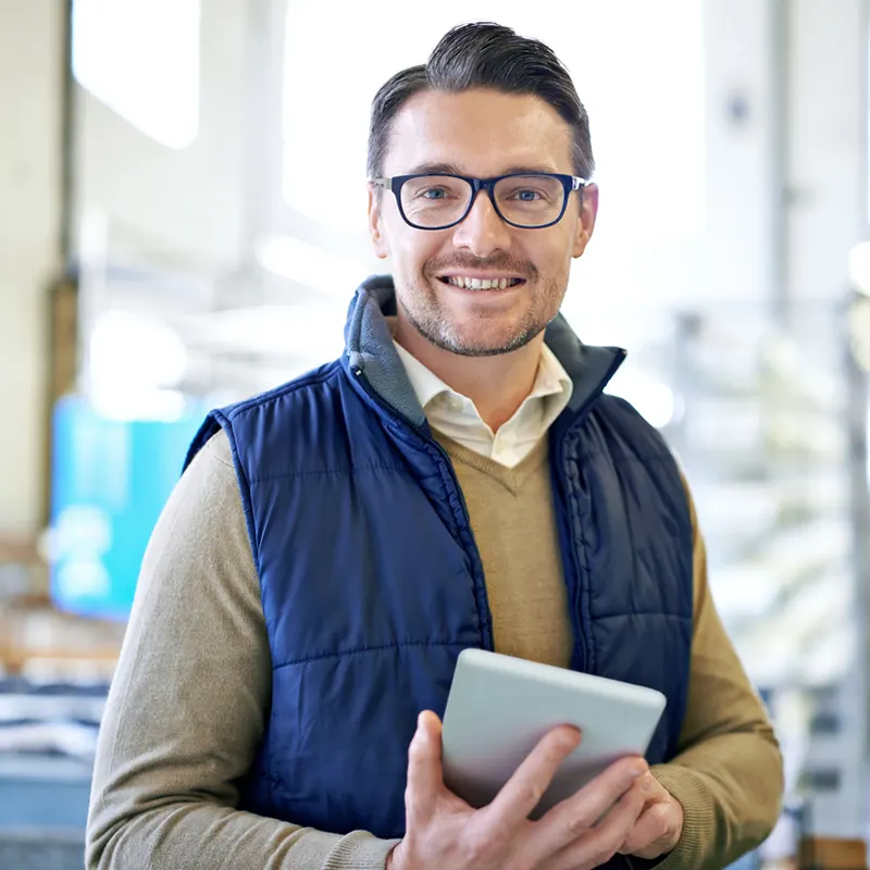 En mann med briller, beige genser og blå vest holder et nettbrett og smiler, mens han står innendørs i et lyst, industrielt miljø i nærheten av en ISO Tank.