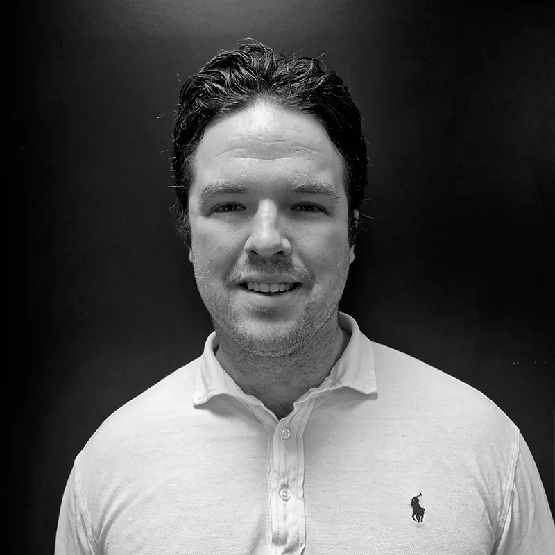 En mørkhåret mann i lys poloskjorte med knapper står mot en ensfarget, mørk bakgrunn, ser inn i kameraet og smiler lett.