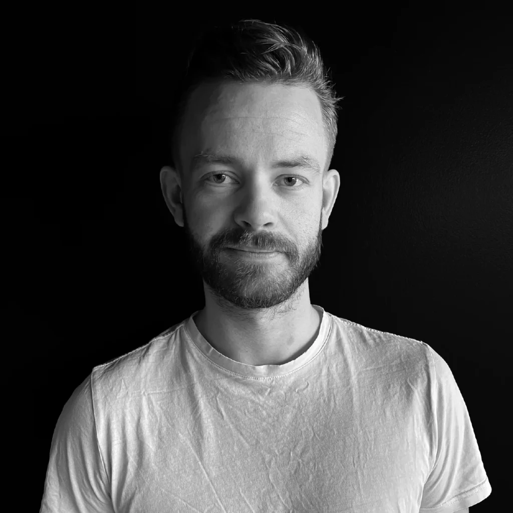 En mann med kort hår og skjegg, iført en vanlig hvit t-skjorte, står mot en mørk bakgrunn og ser direkte inn i kameraet med et nøytralt uttrykk.